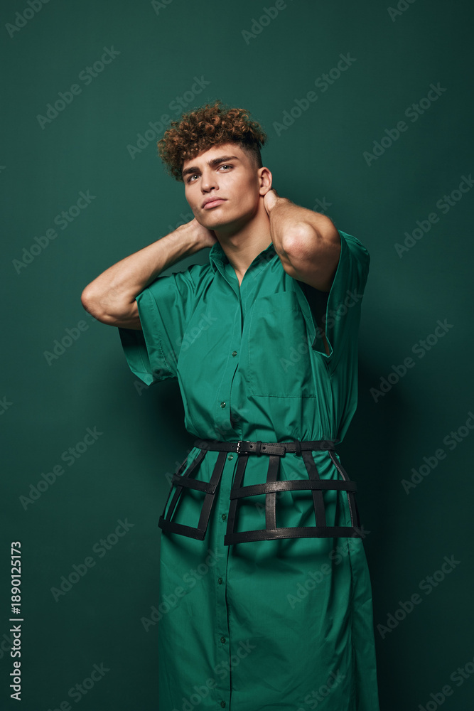 Fototapeta premium Young man with curly hair wearing green dress and black belt posing with hands behind neck against green background in studio. Fashion, style and confident model portrait.