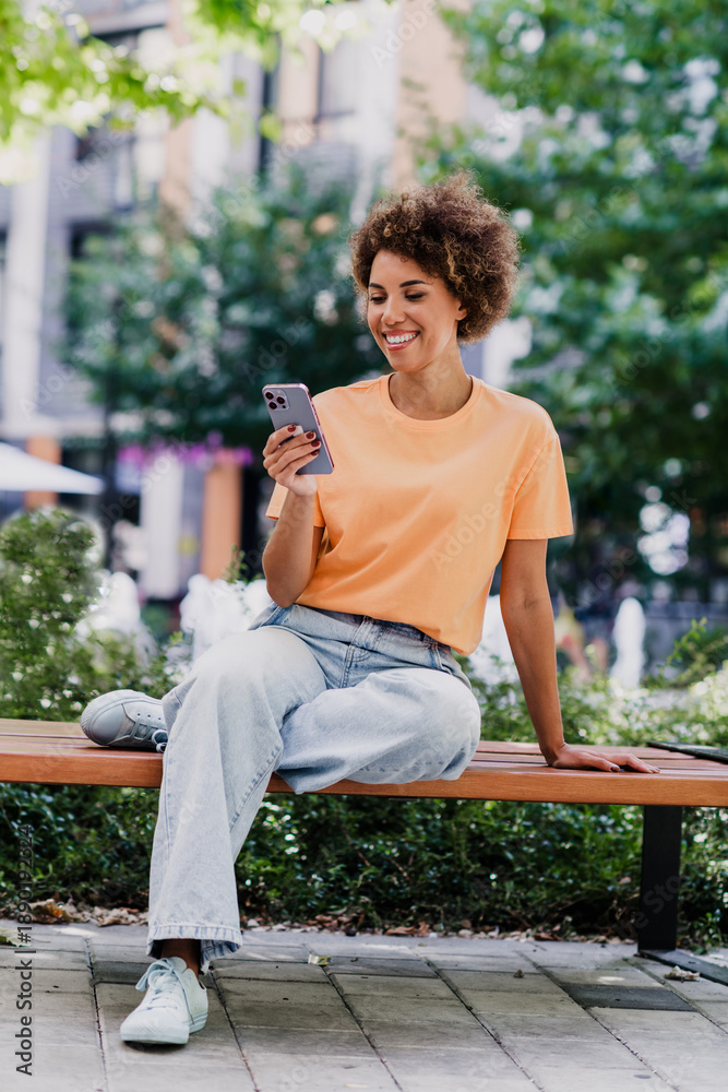 Naklejka premium Relaxed city portrait of an African American woman in a peach shirt sitting on a bench, smiling at her phone in a sunny urban plaza