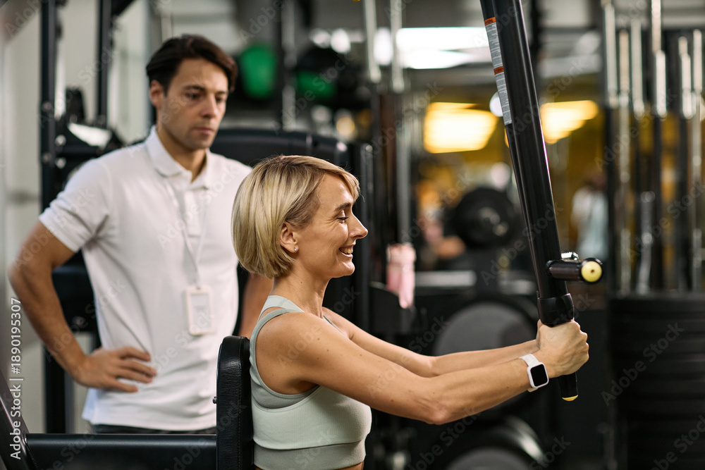 Fototapeta premium Caucasian young adult woman performing strength training exercise on resistance machine under supervision of Caucasian young adult man in physical therapy rehabilitation center