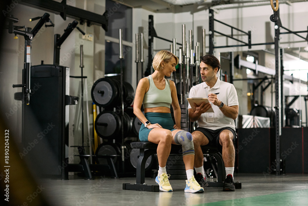 Fototapeta premium Caucasian young adult woman with knee brace sitting on bench talking to Caucasian young adult male physical therapist holding clipboard in rehabilitation gym setting