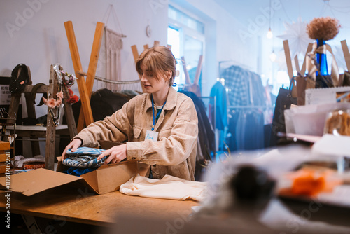 Young Woman Of Caucasian Descent Carefully Packing Clothes Into Boxes At Quaint Store For Future Use