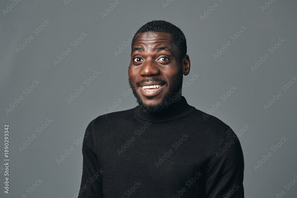Fototapeta premium African man with dark skin wearing black turtleneck sweater posing with surprised expression in creative conceptual visual style on gray background. Studio portrait of young male model.