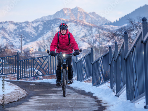 Male cyclist wearing a red jacket and helmet rides an electric bicycle. On a snowy road with mountains in the background in winter in a picturesque outdoor setting