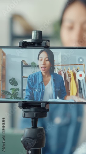 Vertical Screen : Young Asia lady fashion designer using mobile phone receiving purchase order and showing clothes recording video live streaming online at shop. Small business owner, market delivery.