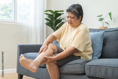 Fotografie Muscle pain or leg pain, suffer asian aged woman, senior massaging brawn leg calf muscle cramps or spasm, older trauma from inflammation of tendon at calves while sitting on sofa