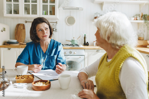 Home visit of female nurse in blue uniform for elderly woman with health issues, social worker collecting complaints before giving medical aid and support both sitting at kitchen table