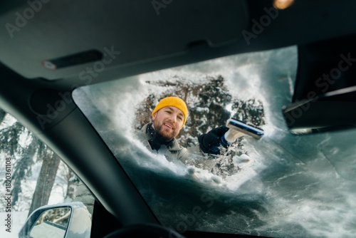 Fotografi Smiling man scraping ice from car windshield in winter