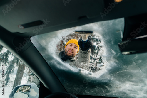 Fotografi Smiling man scraping ice from car windshield in winter