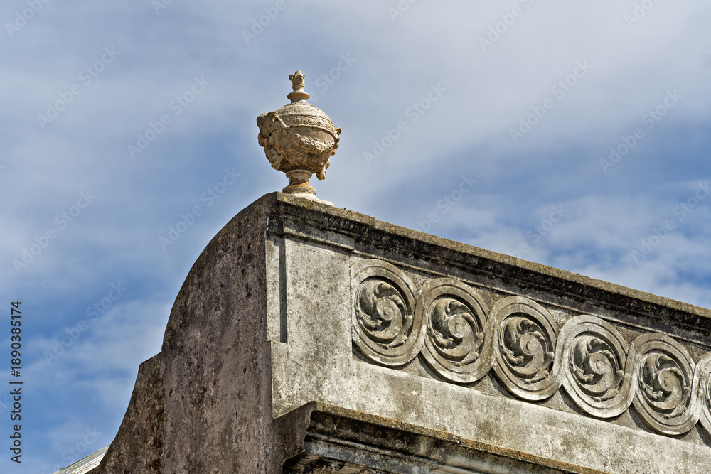 Obraz premium detail in Leirosa, Portugal, showcasing classical stonework with a sculpted urn or finial crowning the upper edge. Beneath it, a frieze features a repeating spiral or volute motif, carved into weather