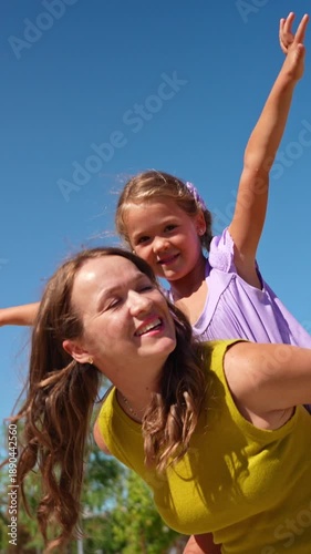 Joyful little girl having fun with her mother on a sunny day. Young woman carrying her cute child on her back against a clear blue sky