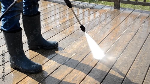 Man in blue jeans and black rubber boots pressure washing dirty wooden deck planks, professional cleaning service