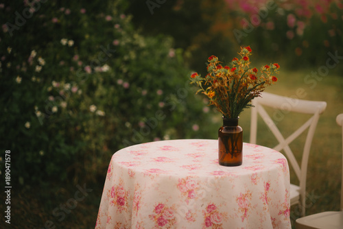 Vintage floral tablecloth brown glass vase wildflower bouquet outdoor garden setting white wooden chair soft natural light summer afternoon rustic decor peaceful atmosphere delicate flowers