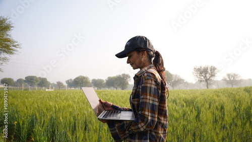 Young woman looking at laptop screen in the middle of wheat field. Business lady inspecting concept, sitting and using notebook. Female farmer wearing cap in middle of crop. A calm, joyful girl