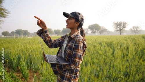 Young woman pointing and using laptop in the wheat field. Business lady inspecting concept, standing and holding notebook, tablet. Female farmer wearing cap in middle of crop. Happy smiling girl