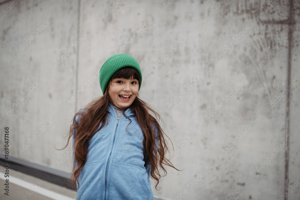 Naklejka premium Outdoor portrait of cute young girl with beanie hat. Girl with long hair standing on playground in the city.