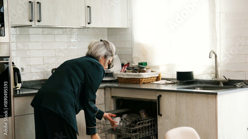 Photos Senior woman doing daily chores, loading dishes into a dishwasher in her kitchen
