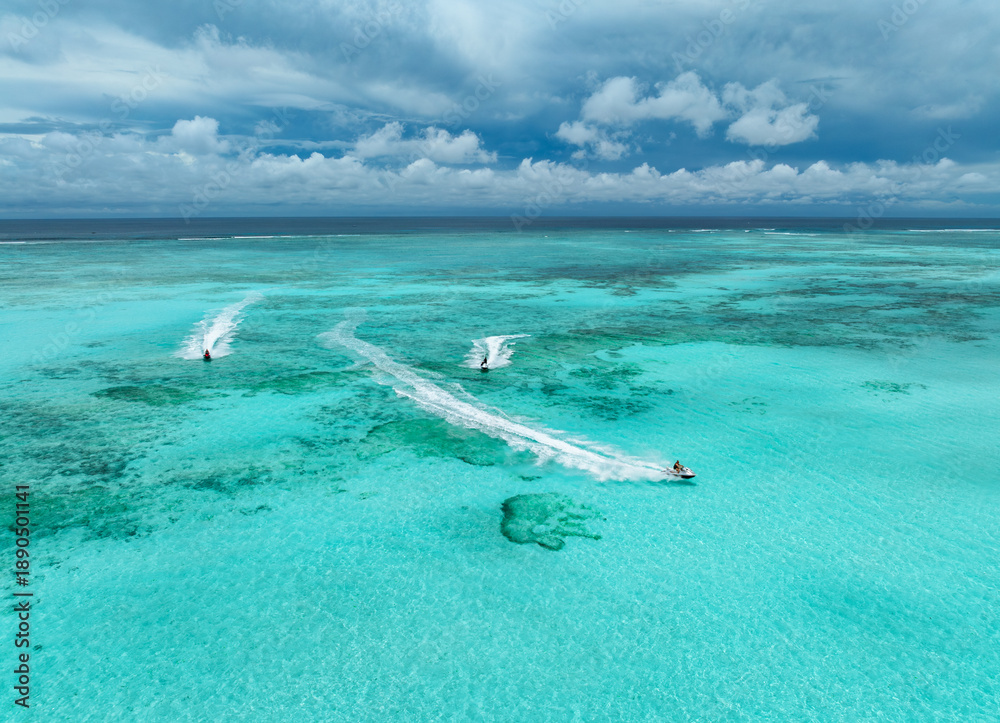 Fototapeta premium Aerial view of jet skis over shallow turquoise sea with reef in Zanzibar. Clear tropical water, watercraft trails, summer vacation, coastal travel and marine leisure activity. Top drone view. Exotic