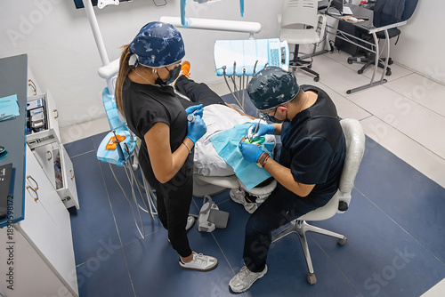 Male dentist and assistant performing dental treatment in a modern clinic, working with professional tools while the patient lies relaxed in the chair during a routine procedure.