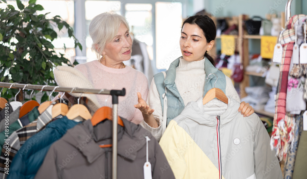 Obraz premium Two interested young and mature women standing in modern clothing shop and looking new coat or jacket