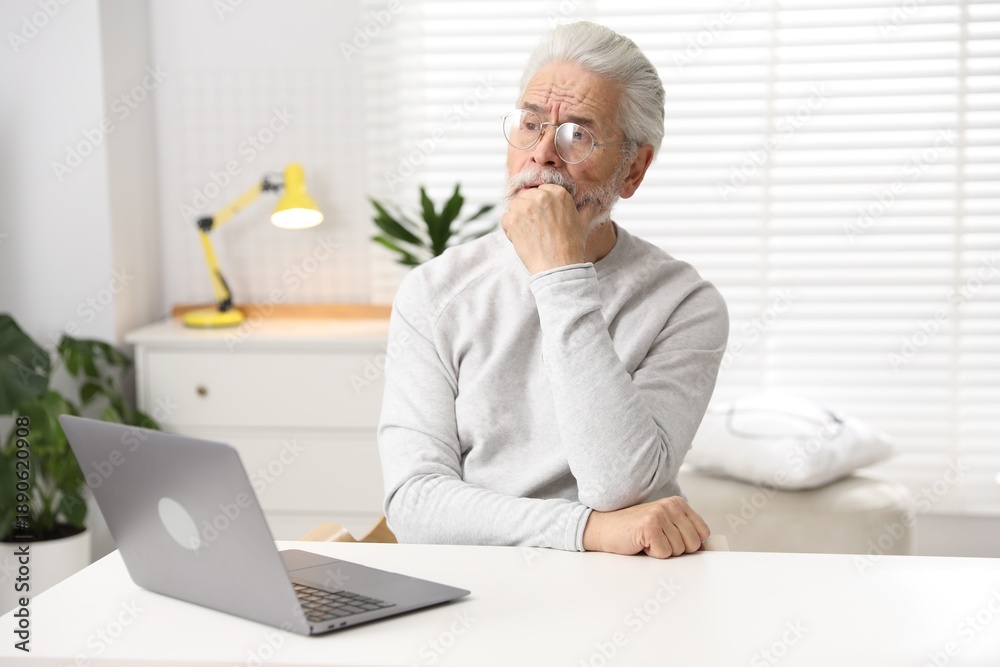 Fototapeta premium Amnesia. Senior man with laptop trying to remember something at white table indoors