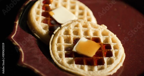 Maple syrup being poured over two toasted frozen waffles with pats of butter on a rustic brown ceramic plate