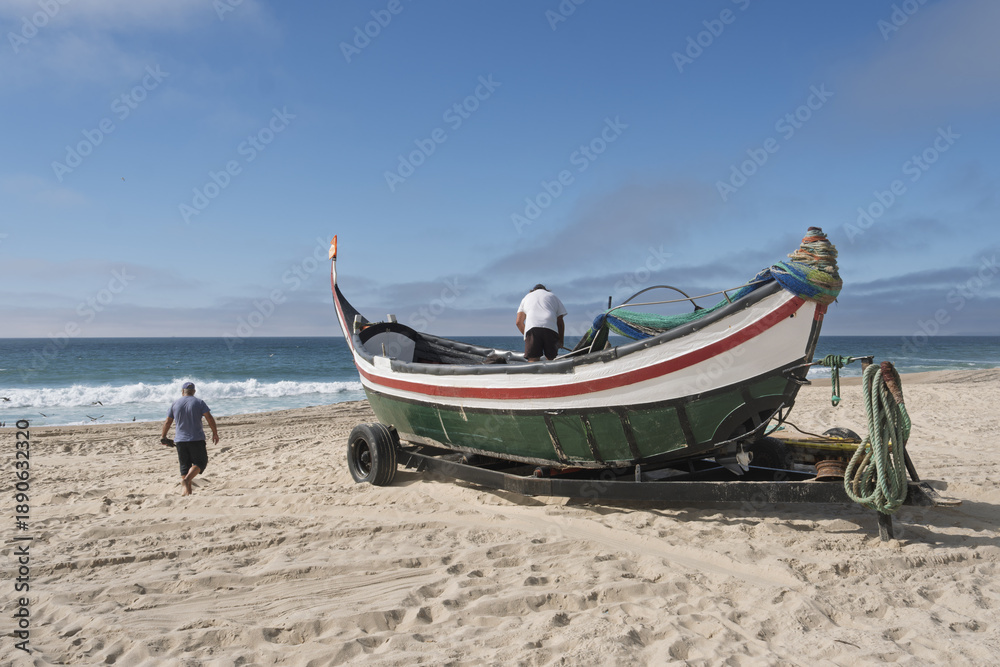 Fototapeta premium Fishing scene on the beach in Leirosa, Portugal, featuring a traditional boat with a curved prow and stern painted in green, white, and red. The boat rests on a trailer 