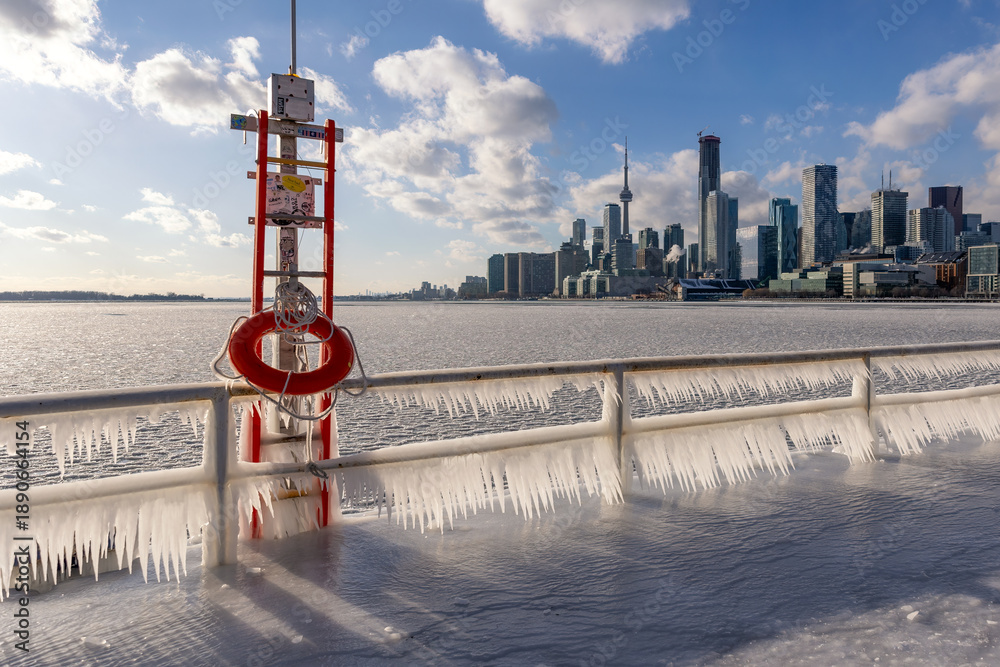 Fototapeta premium Icicles forming on a rail at a frozen pier - Toronto skyline in the background from Polson Pier