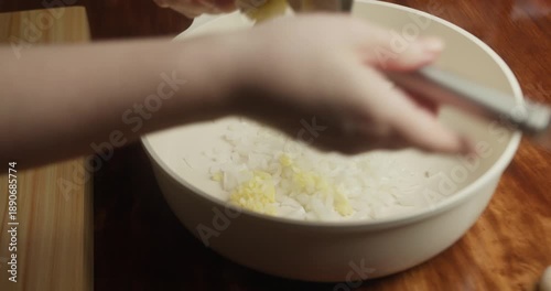 Close-up of a chef pressing fresh garlic into a white bowl with chopped onions using a metal garlic press. Ingredient preparation for homemade cooking in natural light, rustic kitchen atmosphere.