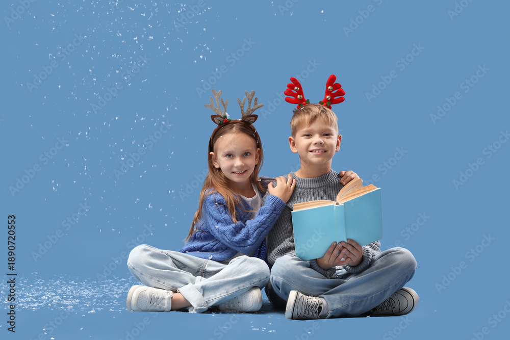 Naklejka premium Cute little children in Christmas reindeer horns with book and snow sitting on blue background