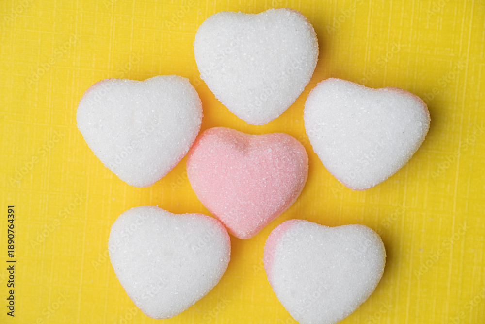 Fototapeta premium A creative arrangement of heart-shaped candies forming a flower shape, with a pink heart in the center surrounded by white hearts on a yellow textured background.