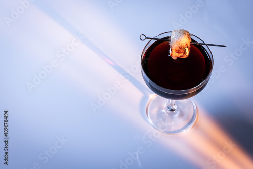 A close-up of a dark cocktail in a coupe glass, illuminated by va warm light in a dark moody background with long shadows.