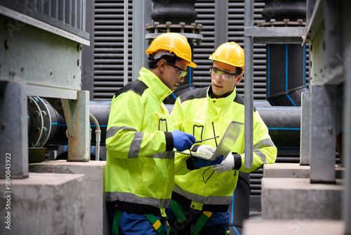 Engineers in high-visibility jackets and hard hats collaborating on site, with modern machinery in background. The workers are using a laptop to review the project