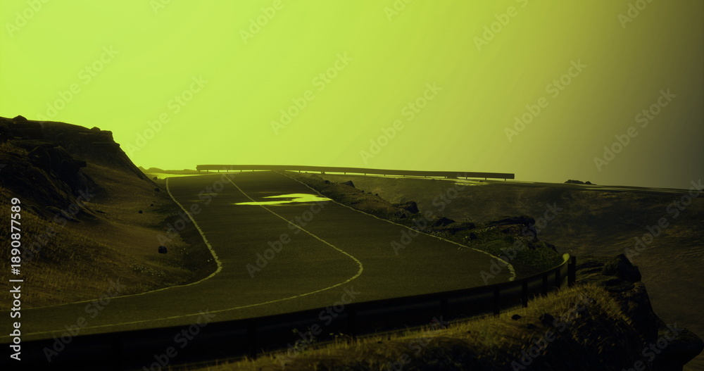 Fototapeta premium A winding road curves through a rugged landscape, bathed in a hazy yellow light as the sun sets. The terrain features rocky outcrops and distant hills, creating a serene atmosphere.