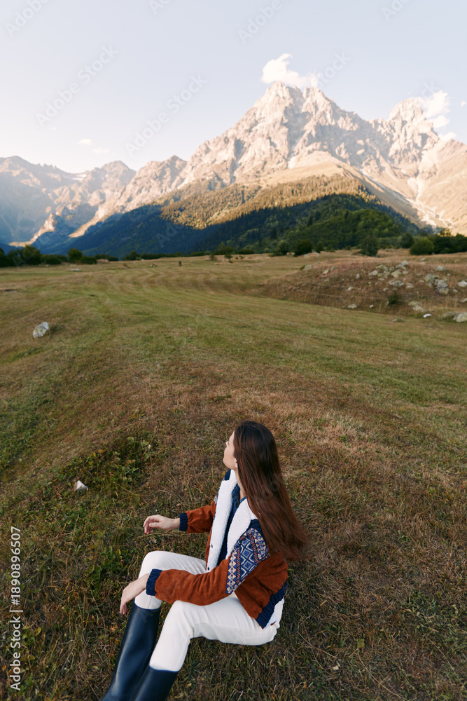 Fototapeta premium woman sitting on meadow facing mountain landscape in nature, autumn travel scene with long hair, cozy jacket and boots, peaceful outdoor relaxation, panoramic alpine valley and hiking vibe