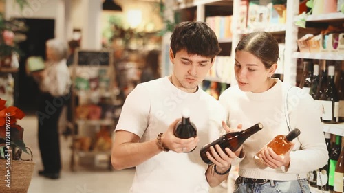 Guy and girl spouses examines bottles of wine near display case, reads product label information. Client compares different options for alcoholic drink, chooses bottle of pink wine.