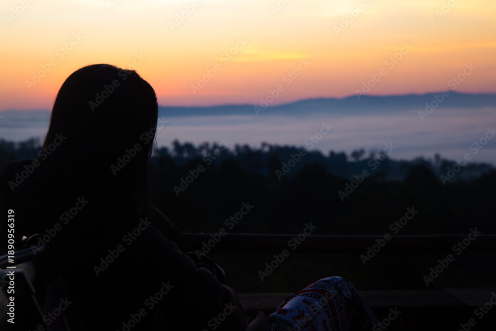 Obraz premium Back view silhouette of woman sitting and watching sea of mist over mountain valley at sunrise, peaceful morning landscape from wooden balcony, concept of freedom, soul searching and slow travel.