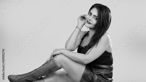 Black and white studio portrait of a confident stylish woman with direct eye contact and a soft smile, seated gracefully in a clean minimal studio environment. Timeless monochrome style, feminine 