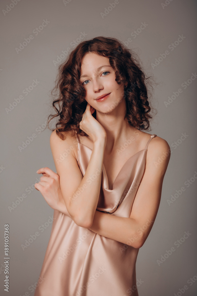 Fototapeta premium The model is a young beautiful brunette girl with curls on a gray background in a pink silk dress