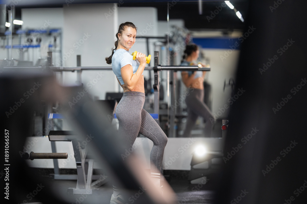 Fototapeta premium Fit woman smiling training bicep with dumbbell in gym