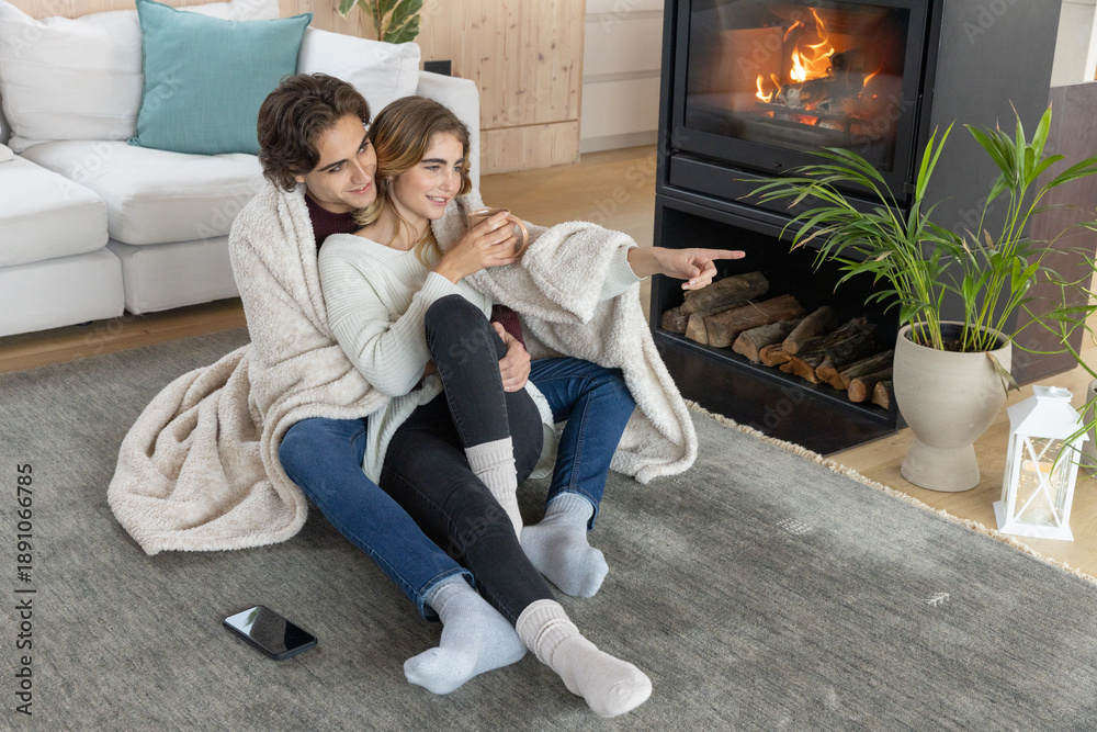 Naklejka premium Couple snuggling together in cream blanket on gray rug in front of fireplace holding white mug