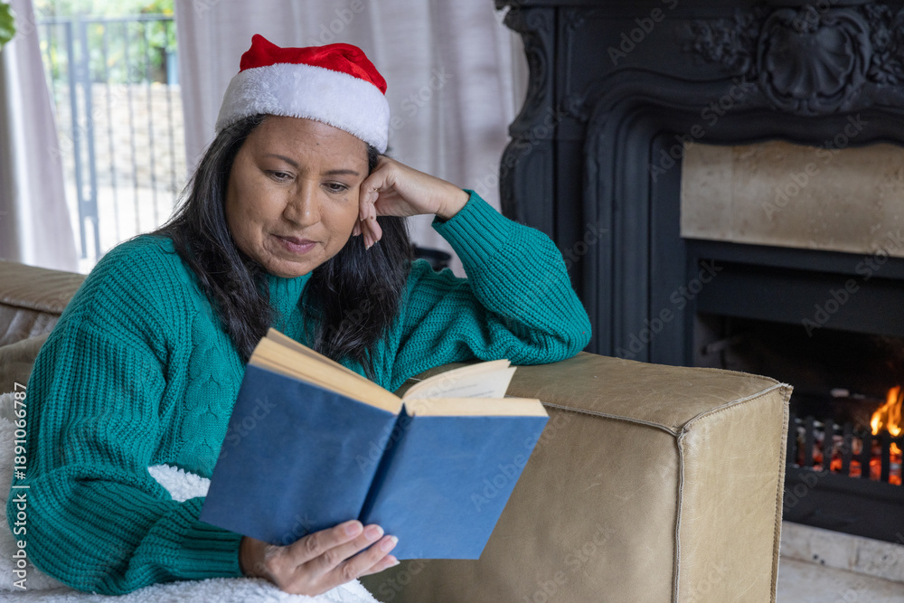 Obraz premium Asian woman sitting in living room on couch in santa hat reading blue book with fireplace