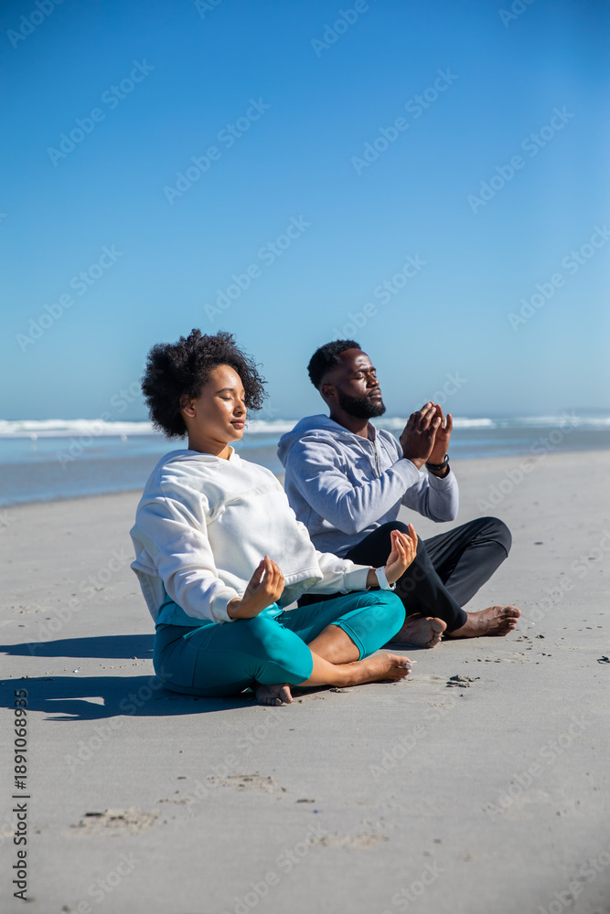 Fototapeta premium African american couple meditating cross-legged at water's edge on ocean beach in activewear