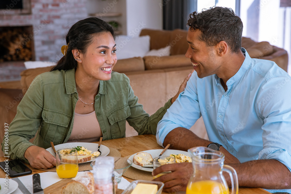 Naklejka premium Diverse couple reaching and smiling while sharing breakfast at home around dining table