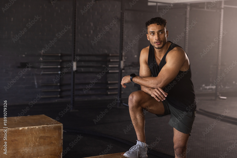 Fototapeta premium Male athlete pausing training kneeling on box beside battle rope wearing tracker in gym, copy space