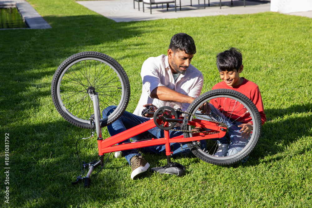 Fototapeta premium Asian father and son kneeling in backyard adjusting red bicycle chain next to helmet and scooter