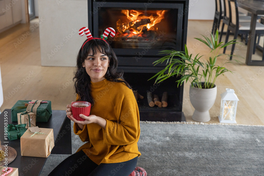 Obraz premium Hispanic woman wearing antler headband holding mug beside fireplace with wrapped gifts