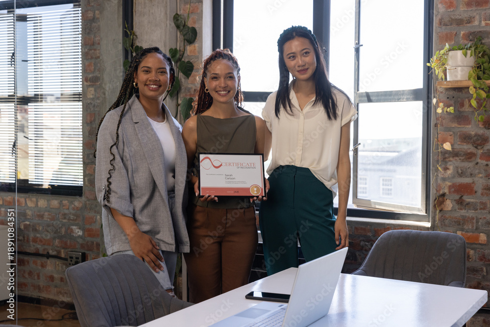 Obraz premium Diverse female coworkers holding framed certificate and smiling in loft office room with laptop