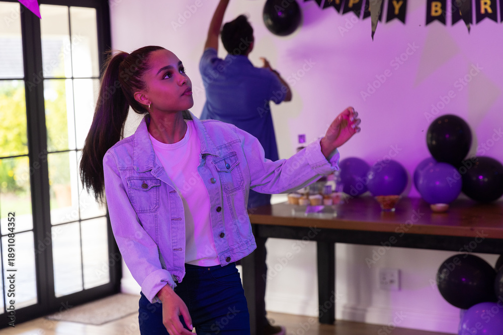 Naklejka premium Diverse friends decorating at home with purple balloons, black-and-gold banner by glass doors