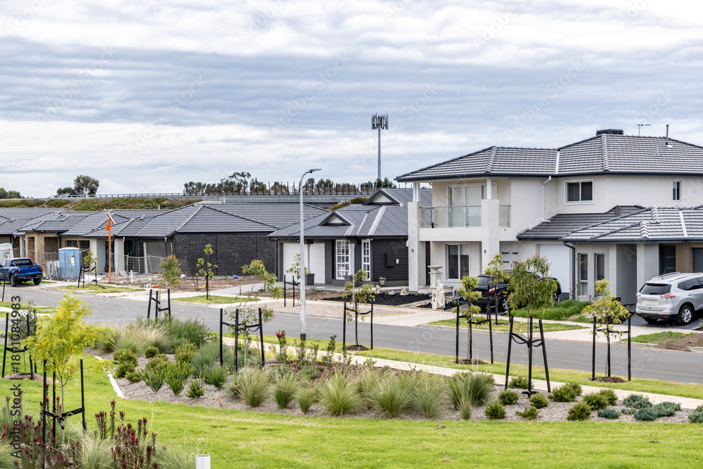 Fototapeta premium Modern suburban street in an outer suburb of Melbourne, Australia, featuring newly built detached houses, landscaped verges, young street trees, and a quiet residential road. Urban growth