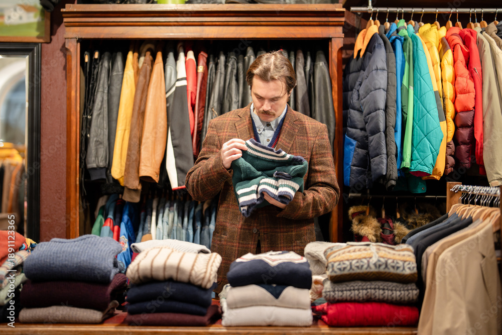 Obraz premium Chic dressed man customer in vintage boutique choosing knitted jumper from display, mustachioed male buyer holding desired apparel item and checking examinees quality of clothing, deciding on trying.